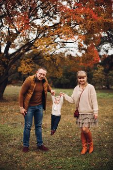 Portrait of attractive young mother and handsome smiling father wearing glasses holding their beautiful lovely baby girl on hands standing against green hedge in autumnal park. They are smiling and looking at camera.