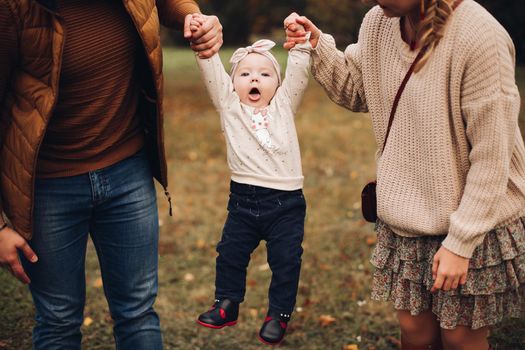 Portrait of attractive young mother and handsome smiling father wearing glasses holding their beautiful lovely baby girl on hands standing against green hedge in autumnal park. They are smiling and looking at camera.