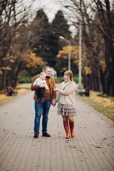 Portrait of attractive young mother and handsome smiling father wearing glasses holding their beautiful lovely baby girl on hands standing against green hedge in autumnal park. They are smiling and looking at camera.