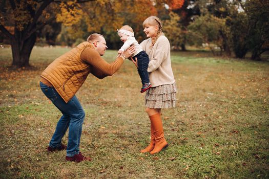 Portrait of attractive young mother and handsome smiling father wearing glasses holding their beautiful lovely baby girl on hands standing against green hedge in autumnal park. They are smiling and looking at camera.