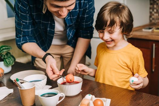 Easter day. Male Father and son painting eggs on wooden background. Family sitting in a kitchen. Preparing for Easter, creative homemade decoration. Child kid boy having fun and painting easter eggs