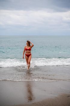 A middle-aged woman with a good figure in a red swimsuit on a pebble beach, running along the shore in the foam of the waves.