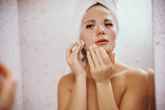 Young beautiful woman using a moisturizing facial mask after taking a bath. Pretty attractive girl in a towel on her head stands in front of a mirror in a home bathroom. Daily hygiene and skin care.