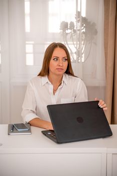 European professional woman is sitting with a laptop at a table in a home office, a positive woman is studying while working on a PC. She is wearing a beige jacket and jeans and is on the phone
