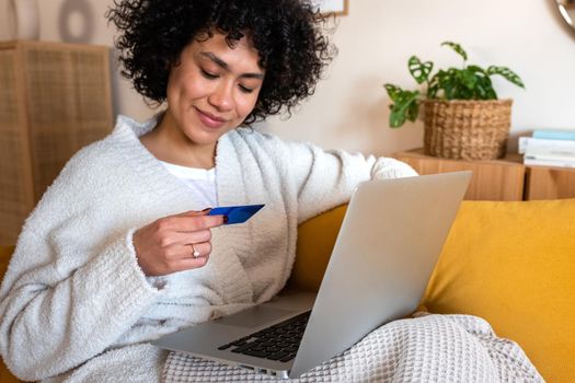 African american woman using laptop and reading credit card information to shop online at home sitting on sofa. Lifestyle and e-commerce concept.