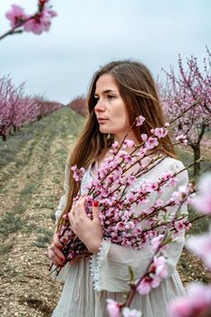 Young beautiful woman in blue dress and long hair is enjoying with blossoming peach trees.