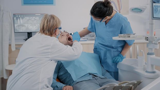 Team of specialists looking at denture after surgical procedure, examining teeth care in dentistry office. Patient with mouth open receiving oral care examination to treat caries.