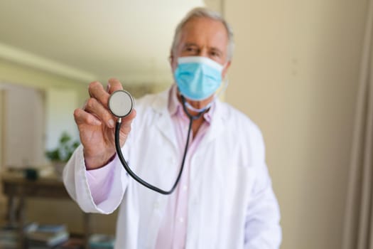 Portrait of senior caucasian male doctor wearing face mask holding stethoscope looking at camera. medicine and healthcare services during covid 19 pandemic concept.