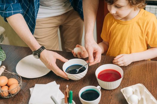 Easter day. Male Father and son painting eggs on wooden background. Family sitting in a kitchen. Preparing for Easter, creative homemade decoration. Child kid boy having fun and painting easter eggs