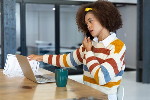 Portrait of mixed race businesswoman sitting at desk with laptop and talking on smartphone. independent creative design business.