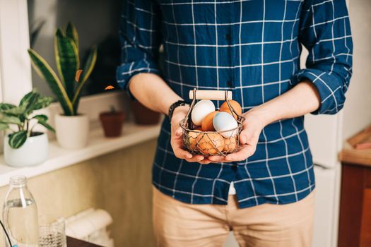 Easter day. Adult man holding basket with eggs on wooden background. Stay in a kitchen with nest. Preparing for Easter, creative homemade decoration.