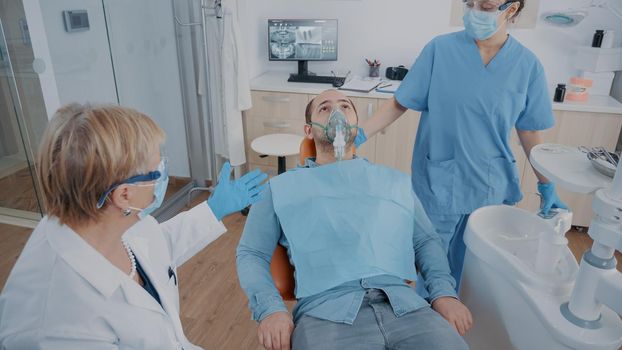 Team of specialists doing anesthesia on patient in dentistry office. Nurse applying oxygen mask on face, giving anesthesic to man before beginning stomatological procedure in cabinet.