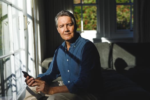 Portrait of happy senior caucasian man in living room sitting on sofa, using smartphone. retirement lifestyle, spending time at home with technology.