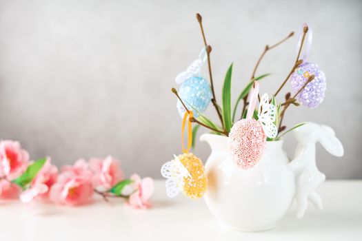 Home interior with easter decor. Vase with willow tree branches with Easter eggs and bunny on white table and background with copy space.