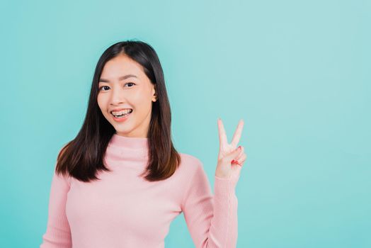 Young Asian beautiful woman smiling wear silicone orthodontic retainers on teeth showing v-sign victory finger isolated on blue background, retaining after removable braces. Dental hygiene concept