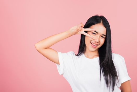 Asian happy portrait beautiful cute young woman teen smile standing showing finger making v-sign victory symbol near eye looking to camera studio shot isolated on pink background with copy space