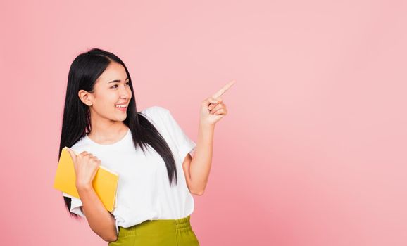 Portrait of happy Asian beautiful young woman confident smiling holding orange book open pointing finger to side copy space, studio shot isolated on pink background, education concept