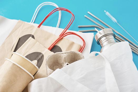 Zero waste concept. Eco friendly reusable items in natural shopping bag. Paper bag and coffee cups, aluminum bottle and metal tubes on blue background