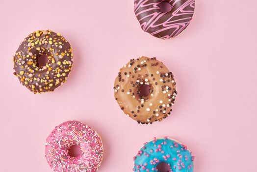 Different types of colorful donats decorated sprinkles and icing on pastel pink background