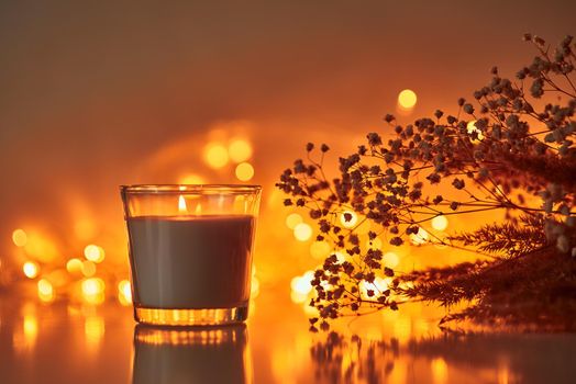 burning candle with dried plant against blurred golden lights on dark background