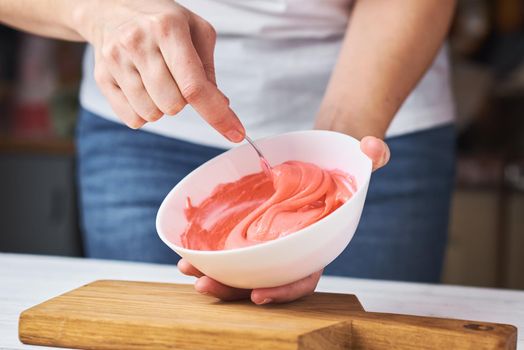 Woman whisking red cream for decorating cookies in bowl on kitchen, closeup