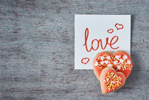 Decorated and glazed heart shape cookies and paper note with inscription LOVE on gray background, top view. Valentines day concept