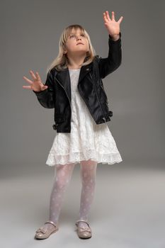 Full length photo of a pretty little blonde girl in a white dress and black leather jacket gesticulating while posing standing over a gray background. Copy space. Sincere emotions concept.