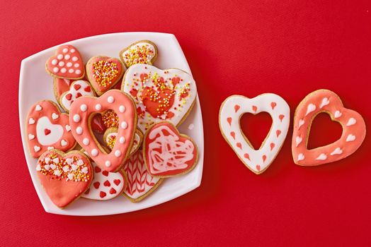 Decorated heart shape cookies in white plate and two cookies on red background, top view. Valentines Day food concept