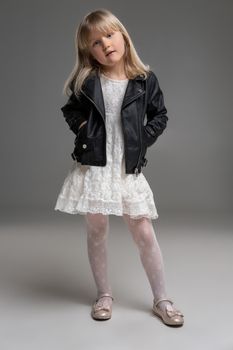 Full length photo of a goegeous little blonde girl in a white dress and black leather jacket looking at the camera and posing standing over a gray background. Copy space. Sincere emotions concept.