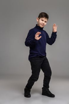 Full length portrait of a nice kid in a dark clothes and black boots gesticulating and looking at the camera while posing at studio as a fashion model. Photo of a schoolboy over a gray background. Copy space. Emotions concept.