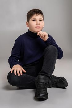 Full length portrait of a nice boy in a dark clothes and black boots sitting on the floor and looking at the camera while posing at studio as a fashion model. Photo of a schoolboy over a gray background. Copy space. Emotions concept.