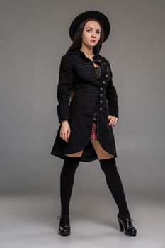 Full length portrait of an attractive woman looking up and posing at studio against a gray background. She is weared in a checkered dress, black coat, hat, stockings and boots. Fashion shot. Sincere emotions concept.