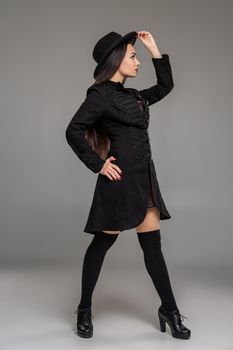 Full length portrait of an attractive professional model posing sideways at studio against a gray background. She is weared in a checkered dress, black coat, hat, stockings and boots. Fashion shot. Sincere emotions concept.