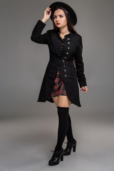 Full length portrait of a beautiful woman looking away while posing at studio against a gray background. She is weared in a checkered dress, black coat, hat, stockings and boots. Fashion shot. Sincere emotions concept.