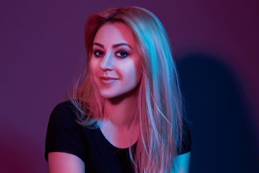 Close-up portrait of a beautiful model with fresh evening makeup and long blond hair, wearing in a black t-shirt. She is posing in a studio against a colorful background and looking at the camera.