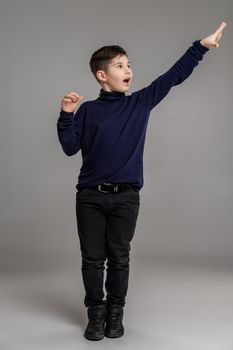 Full length portrait of a nice little man in a dark clothes and black boots gesticulating and looking away while posing at studio as a fashion model. Photo of a schoolboy over a gray background. Copy space. Emotions concept.