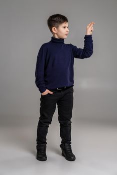 Full length portrait of a nice fellow in a dark clothes and black boots acting like holding something while posing at studio as a fashion model. Photo of a schoolboy over a gray background. Copy space. Emotions concept.
