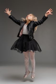 Full length photo of a charming little blonde child in a white dress and black leather jacket jumping while posing standing over a gray background. Copy space. Sincere emotions concept.