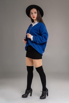 Full length portrait of a gorgeous woman looking away while posing at studio against a gray background. She is weared in a oversize blue blouse, black hat, skirt, stockings and boots. Fashion shot. Sincere emotions concept.