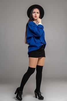 Full length portrait of an alluring woman posing at studio against a gray background. She is weared in a oversize blue blouse, black hat, skirt, stockings and boots. Fashion shot. Sincere emotions concept.