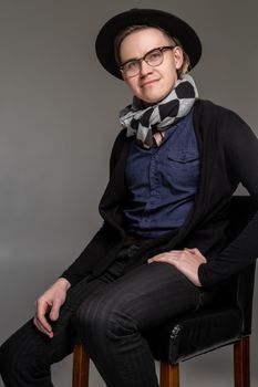 Pensive brunet man in a casual dark clothes, stylish scarf, glases and black hat is looking at the camera, smiling and sitting on the bar chair while posing over a gray background. Copy space, close-up. Emotions concept.
