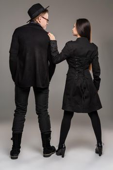 Young loving couple. Studio shot of a beautiful young female and handsome male. They are in black outlet clothes and stylish hat posing standing back to the camera over a gray studio background. Fashion shot. Sincere emotions concept. Copy space.