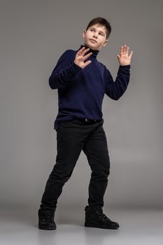 Full length portrait of a nice child in a dark clothes and black boots gesticulating and posing at studio as a fashion model. Photo of a schoolboy over a gray background. Copy space. Emotions concept.