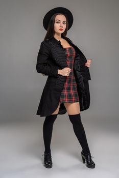 Full length portrait of a charming girl looking at the camera and posing at studio against a gray background. She is weared in a checkered dress, black coat, hat, stockings and boots. Fashion shot. Sincere emotions concept.