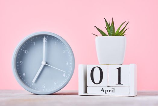 Wooden blocks calendar with date 1 april, alarm clock and plant on pink background