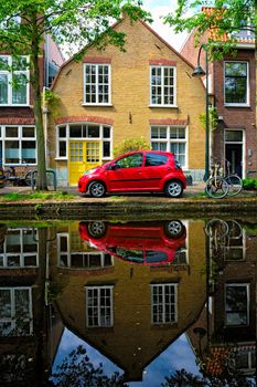 Red car on canal embankment in street of Delft with reflection and bicycle. Delft, Netherlands