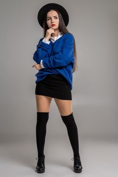 Full length portrait of an alluring female folded her hands and looking away while posing at studio against a gray background. She is weared in a oversize blue blouse, black hat, skirt, stockings and boots. Fashion shot. Sincere emotions concept.