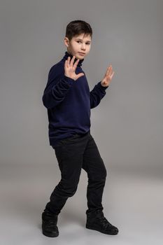 Full length portrait of a nice guy in a dark clothes and black boots has raised his hands up and looking at the camera while posing at studio as a fashion model. Photo of a schoolboy over a gray background. Copy space. Emotions concept.