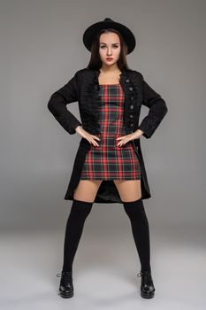 Full length portrait of a charming lady looking straight and posing at studio against a gray background. She is weared in a checkered dress, black coat, hat, stockings and boots. Fashion shot. Sincere emotions concept.