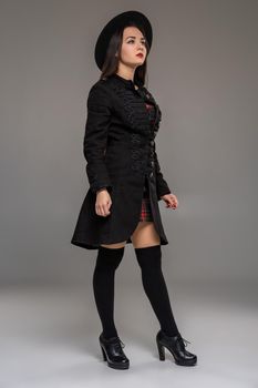 Full length portrait of a charming woman looking up and posing sideways at studio against a gray background. She is weared in a checkered dress, black coat, hat, stockings and boots. Fashion shot. Sincere emotions concept.